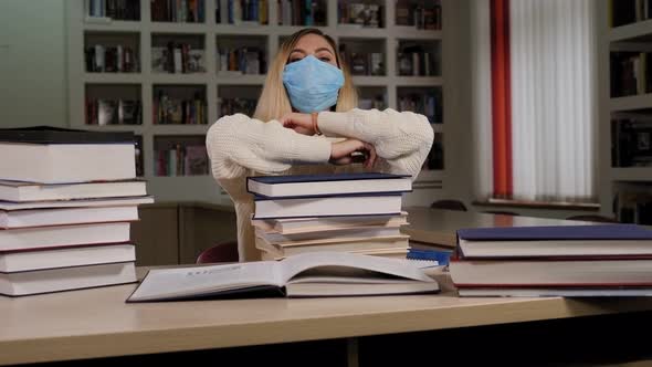 Young Girl Student in Medical Mask Lies on a Large Pile of Books in the Library alt