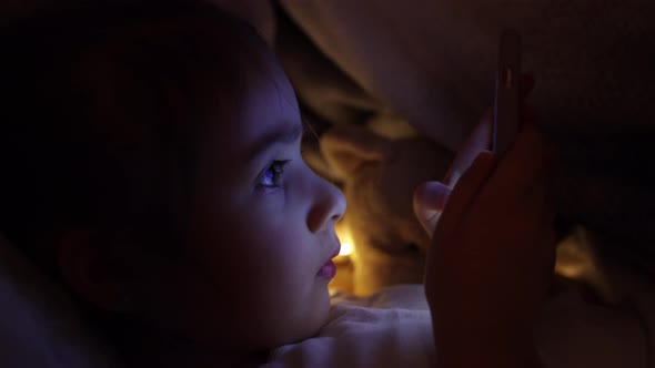 Cute baby girl uses phone in bed at night instead of sleeping close up.