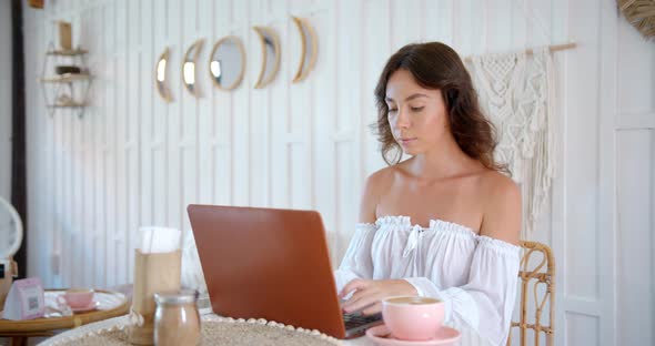 Young Woman Works at White Cozy Cafe She Drinks Coffee and Typing on Her Lap Top Computer alt