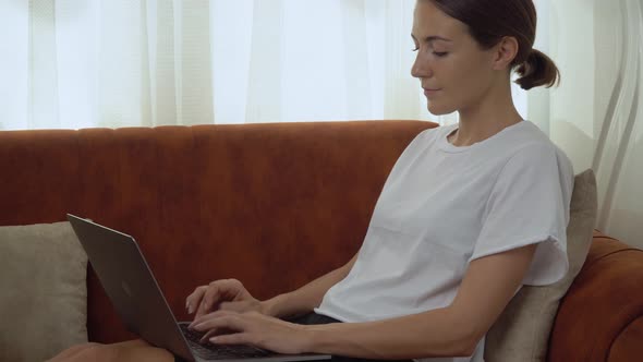A Woman Works at a Laptop on the Couch Typing on the Keyboard
