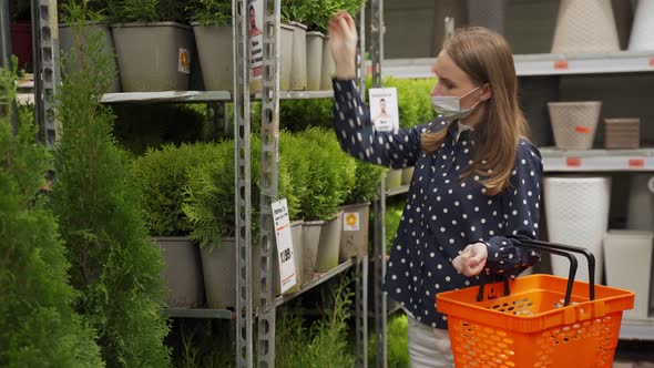 Young Woman in a Protective Mask Chooses and Buys a Tuya at a Garden Center in Early Spring alt