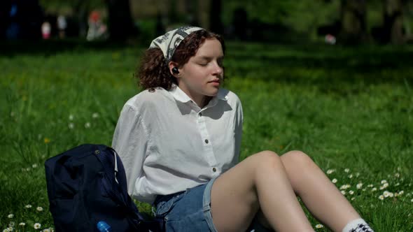 Teenage Girl Sits on the Green Grass in the Park and Listens to Music Eyes Closed Through Earphone