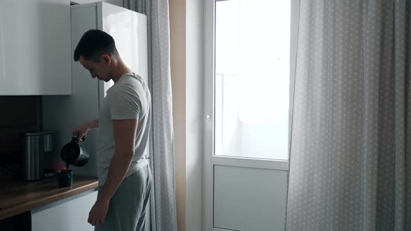 Handsome Young Man Pours Coffee From Coffee Maker and Walks Up to Window in Kitchen alt