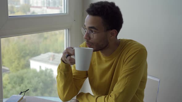 A Young African Man is Drinking Coffee While Sitting By the Window alt