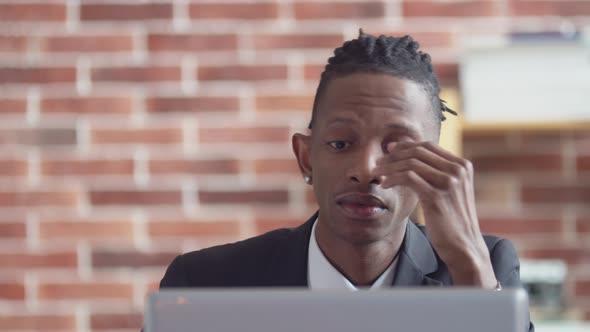 Black Tired Man in a Suit Takes Off Glasses and Rubs Eyes with Hands While Sitting at an Office alt