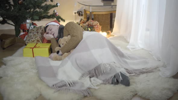 Wide Shot of Happy Young Man Sleeping on Gift Boxes at Christmas Tree at Home alt