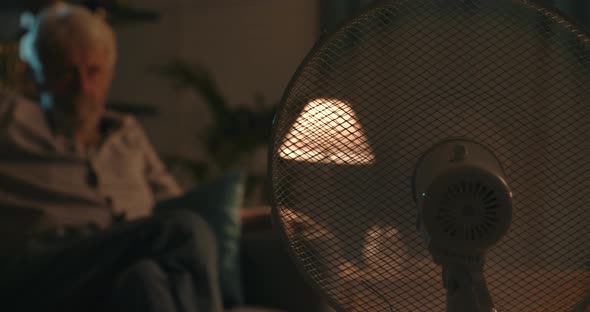 Senior man watching TV at home on a summer night, fan in the foreground