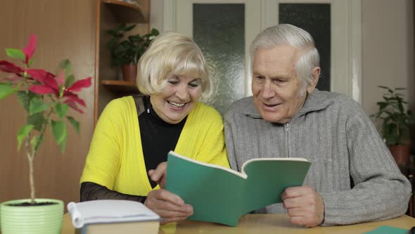 Happy Old Grandparents Couple Sit on Table at Home Enjoy Reading Book Together alt