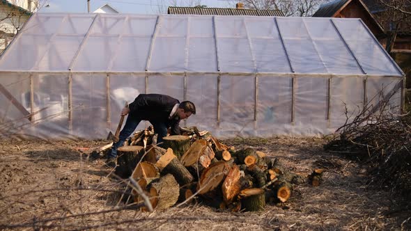 A young man is chopping wood with an axe in the garden near the greenhouse.