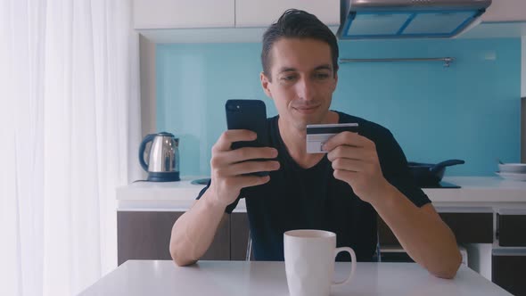 Young Handsome Man Sitting at Table Using Smart Phone for Online Shopping with Credit Card at Home alt