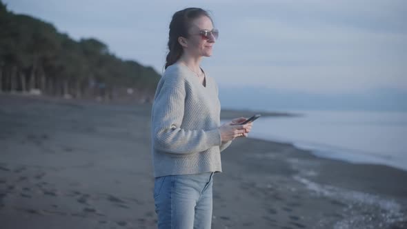 Young Attractive Relaxed Woman Enjoys Sunrise on Sea Coast and Holds Smartphone