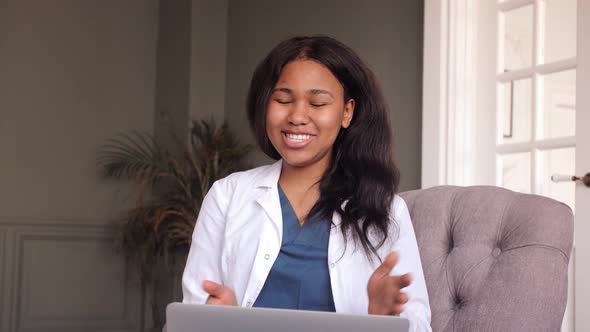 Friendly Female Black Clinician in White Gown Using Laptop for Remote Patient Consultation alt
