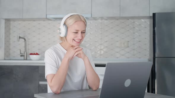Young Blonde Woman in Headphones Starts Video Call Sitting at Laptop in Kitchen