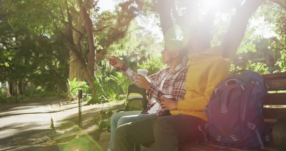 Two happy mixed race male friends sitting and talking in park with backpacks alt