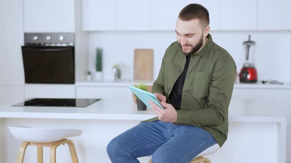 Absorbed Young Bearded Man Messaging Online or Surfing Social Media on Tablet Smiling alt