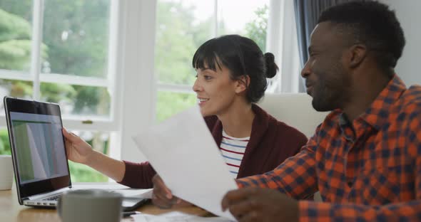 Happy diverse couple sitting at table talking and working with laptop alt