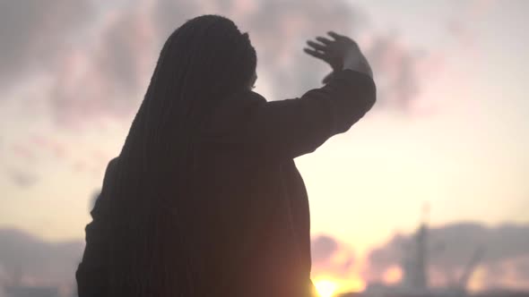 Serene black woman outstretching arm against bright setting sun alt