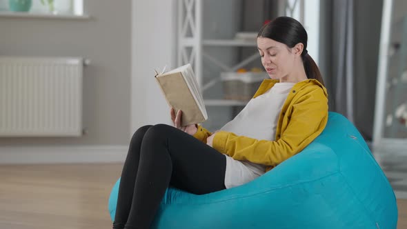 Side View Smiling Pregnant Woman Reading Book Stroking Belly Sitting on Bag Chair Indoors alt