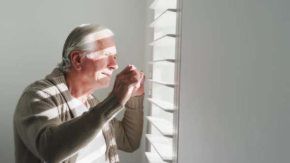 Side view of retired senior man in cardigan sweater looking out through window blinds at home alt