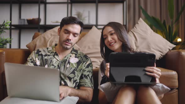 Young couple using tablet and laptop for work together on sofa in living room at home