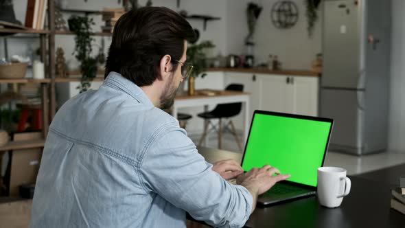 Over the shoulder shot of a young Caucasian man sitting, working from home on a laptop, green screen