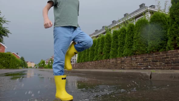 Little boy wearing yellow rubber boots jumping in puddle of water on rainy summer day in small town