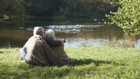 Elderly Gray Haired Couple Siting on the Bench Near the River on Autumn Day Covered with Blanket alt
