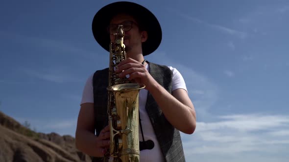 Musician Is Dressed in Hat and Waistcoat and Playing Saxophone at Nature at Summer Day alt