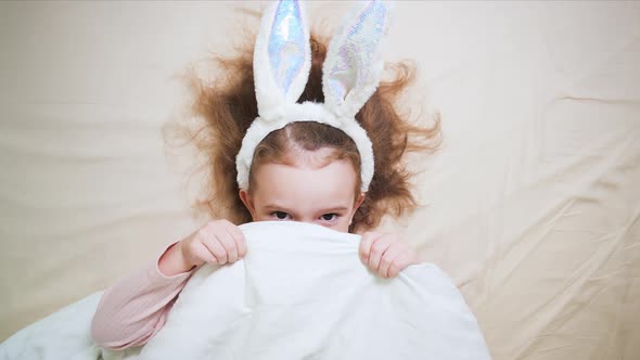 A Cute Little Girl with Bunny Ears is Lying in Bed and Hiding alt