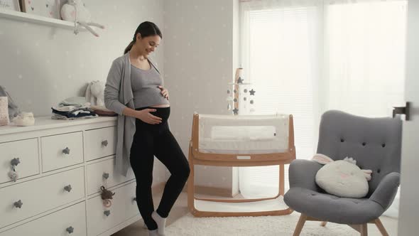 Caucasian woman in advanced pregnancy standing in the baby's room next to the crib. alt