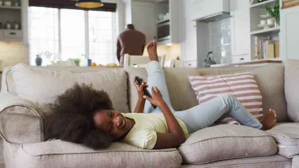 African american daughter lying on couch laughing at tv with father in background alt