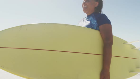 Happy senior african american woman walking with surfboard on sunny beach alt