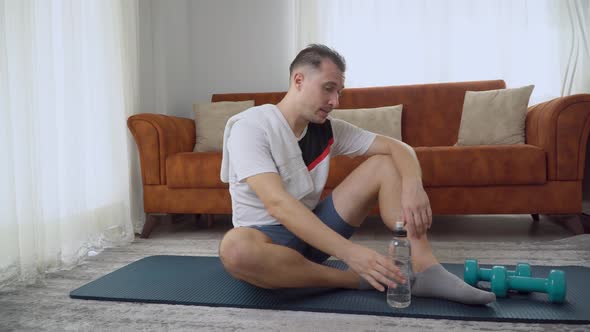 A Man Drinks Water After a Workout at Home Sitting on a Mat with Dumbbells