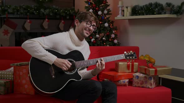 Man is playing Christmas song on guitar near decorated Christmas tree.