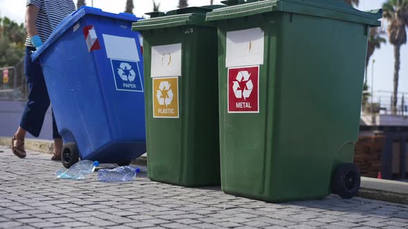 Unrecognizable Woman Rolling Paper Container to Plastic and Metal Waste Bins on Coastal Pavement alt
