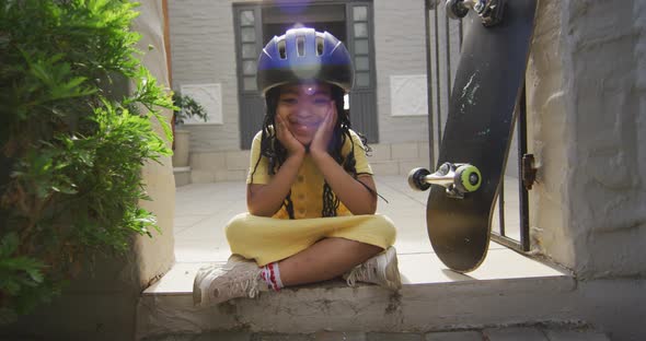 Happy african american girl wearing helmet sitting on stairs with skateboard and looking at camera alt