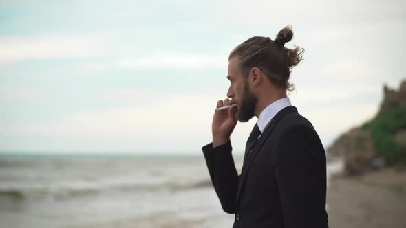 Young Guy in Business Suit with Tied Hair Thoughtfully Watching in Distance at Sea and Smoking alt