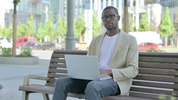 African Man with Laptop Looking at Camera While Sitting Outdoor on Bench alt