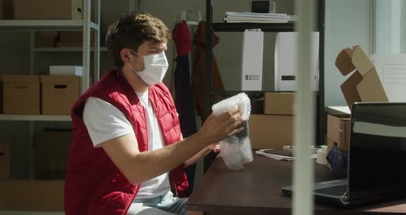 Man Wearing Red Vest Protective Mask Employee of Warehouse Packing Parcel Into Delivery Box and alt