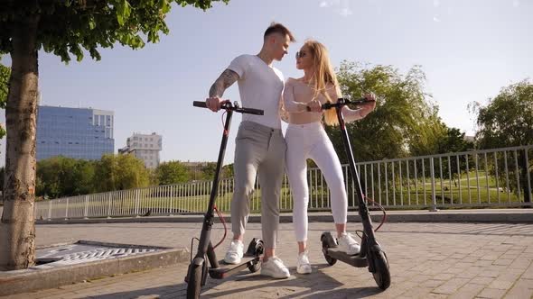 A Couple of Teenagers Hug in a Park in the Summer, Next To an Electric Scooter. alt