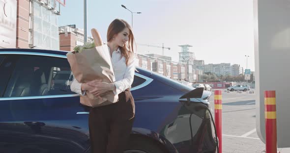 Beautiful Business Woman Stands Near Her Electric Car With Groceries alt