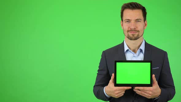 A Young Businessman Smiles at the Camera and Holds Out a Tablet with Green Screen alt