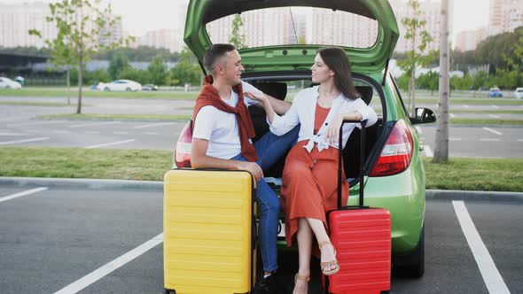 A Happy Young Couple are Sitting in the Trunk of a Car Leaning on Colorful Travel Suitcases