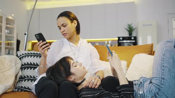Two Girls are Relaxing on the Couch Each Looking at Her Smartphone alt