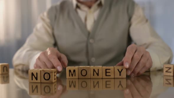 Mature Man Making Words No Money of Wooden Cubes on Table, Pension Age Poverty alt