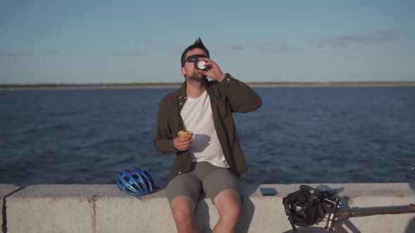 Male Cyclist Eats Sandwich and Drinks Coffee to Go While Sitting on Promenade By Sea alt