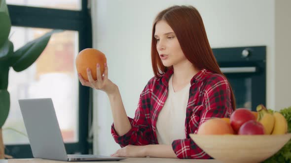 Caucasian Young Woman Food Blogger Redhead Nutritionist Sits at Kitchen Shows at Webcam Grapefruit alt