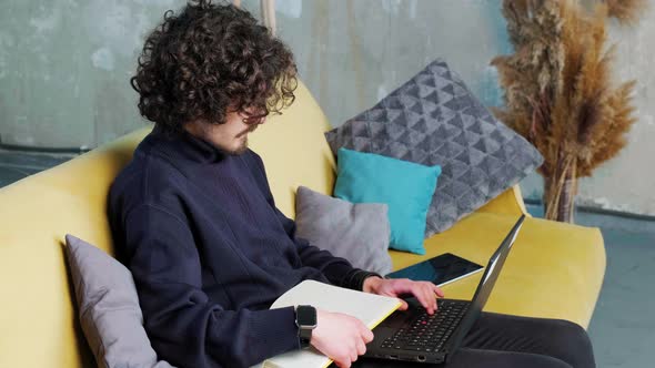 Young Man is Browsing at His Laptop and Writing Notice on His Paper Notebook alt