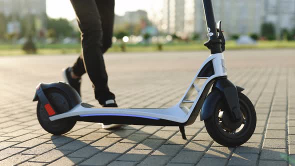 Shot on Legs Young Hipster Man in Black Sneakers Ride on Electric Mobile Scooter at Sunset alt