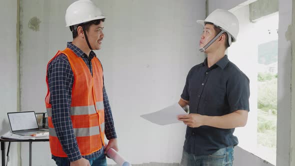 Two Asain confident engineers discussing blueprint while standing at construction site
